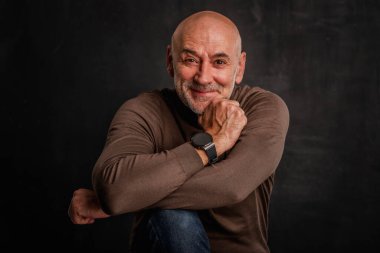 Siyah arka planda kameraya gülümseyen olgun bir adamın portresi. Balıkçı yaka kazak giyen kendinden emin bir erkek..