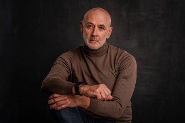 Tecrit edilmiş siyah arka planda olgun bir adamın portresi. Kendine güvenen erkek balıkçı yaka kazak giyiyor ve ne yazık ki kameraya bakıyor. Boşluğu kopyala. 