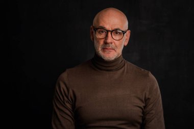 Tecrit edilmiş siyah arka planda olgun bir adamın portresi. Ciddi yüzlü, gözlüklü ve balıkçı yaka kazaklı bir erkek. Boşluğu kopyala. 