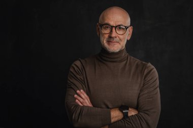 Tecrit edilmiş siyah arka planda olgun bir adamın portresi. Gözlüklü ve balıkçı yaka kazaklı kendinden emin bir erkek. Boşluğu kopyala. 