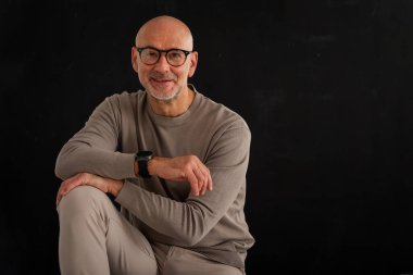 Siyah arka planda kameraya gülümseyen gözlüklü olgun bir adamın portresi. Balıkçı yaka kazak giyen kendinden emin bir erkek..