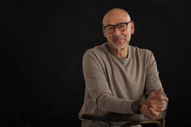 Siyah arka planda kameraya gülümseyen gözlüklü olgun bir adamın portresi. Balıkçı yaka kazak giyen kendinden emin bir erkek..