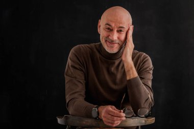 Siyah arka planda kameraya gülümseyen olgun bir adamın portresi. Balıkçı yaka kazak giyen kendinden emin bir erkek..