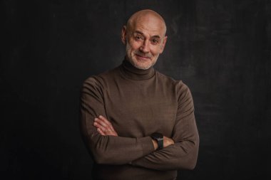 Siyah arka planda kameraya gülümseyen olgun bir adamın portresi. Balıkçı yaka kazak giyen kendinden emin bir erkek..