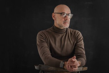 Gözlüklü olgun bir adamın portresi izole edilmiş siyah arka plana dikkatlice bakıyor. Balıkçı yaka kazak giyen kendinden emin bir erkek..