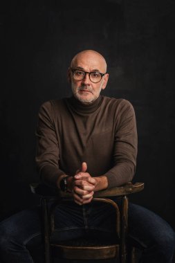 Orta yaşlı bir adam düşünceli görünüyor ve siyah arka planda oturuyor. Kel ve sakallı bir adam, izole bir arka planda günlük kıyafetler ve gözlük takıyor. Boşluğu kopyala. 