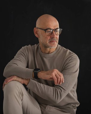Orta yaşlı bir adam düşünceli görünüyor ve siyah arka planda oturuyor. Kel ve sakallı bir adam, izole bir arka planda günlük kıyafetler ve gözlük takıyor. Boşluğu kopyala. 
