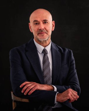 Orta yaşlı bir adamın takım elbise giyip izole edilmiş karanlık arka planda oturduğu bir fotoğraf. Boşluğu kopyala.