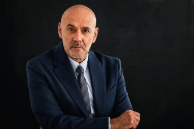 Kızgın suratlı, takım elbiseli ve izole edilmiş karanlık arka planda oturan bir adamın stüdyo fotoğrafı. Boşluğu kopyala.
