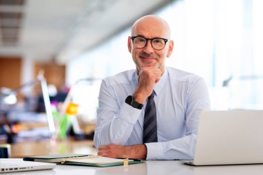 Ofiste oturan orta yaşlı bir işadamı ve not defterini iş için kullanıyor. Gömlek ve kravat takan ve kameraya bakan kendine güvenen profesyonel bir adam..