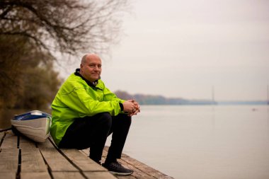 Orta yaşlı bir adam kano yaptıktan sonra iskelede oturuyor. Aktif sporcu kuru takım elbise giyiyor ve kameraya bakıyor.. 