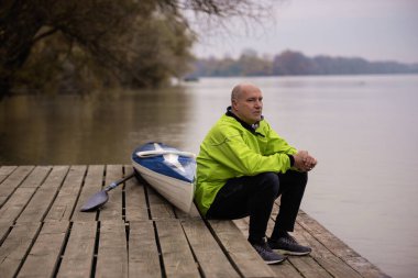 İskelede oturan ve kanoya binmeye hazır orta yaşlı bir adam. Aktif sporcu kuru takım elbise giyiyor ve başka tarafa bakıyor.. 
