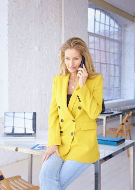 Sarı ceket ve kot pantolon giyen ve ofiste duran çekici orta yaşlı kadın. Profesyonel kadın akıllı telefonunu kullanıyor ve telefonda biriyle konuşuyor..