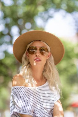 Sokakta otururken güneş gözlüğü ve şapka takan çekici sarı saçlı kızın yakın plan portresi.. 