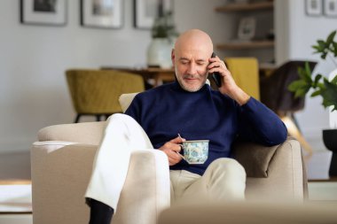 Orta yaşlı bir adamın portresi cep telefonu kullanıyor ve evdeki koltukta dinlenirken telefonla konuşuyor. Balıkçı yaka kazak giyen ve çay içen mutlu bir erkek..