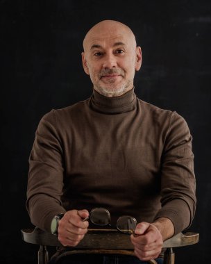 Siyah arka planda kameraya gülümseyen olgun bir adamın portresi. Balıkçı yaka kazak giyen kendinden emin bir erkek..