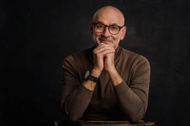 Siyah arka planda kameraya gülümseyen olgun bir adamın portresi. Balıkçı yaka kazak giyen kendinden emin bir erkek..