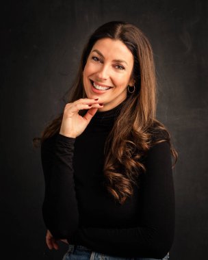 Studio portrait of an attractive middle aged woman with toothy smile wearing turtleneck sweater while sitting at isolated dark background. Copy space.