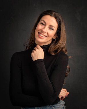 Studio portrait of an attractive middle aged woman with toothy smile wearing turtleneck sweater while sitting at isolated dark background. Copy space.