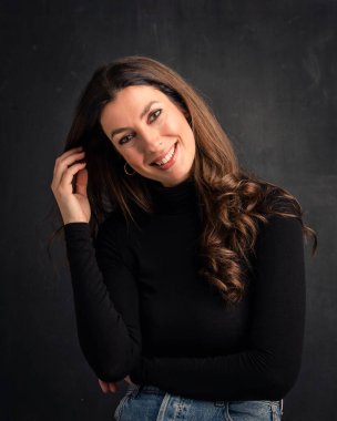 Studio portrait of an attractive middle aged woman with toothy smile wearing turtleneck sweater while sitting at isolated dark background. Copy space.