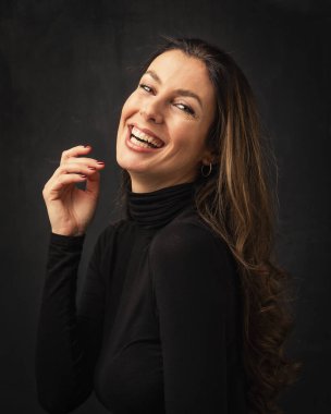Studio portrait of an attractive middle aged woman with toothy smile wearing turtleneck sweater while sitting at isolated dark background. Copy space.