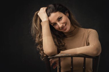 Studio portrait of an attractive middle aged woman with toothy smile wearing turtleneck sweater while sitting at isolated dark background. Copy space.