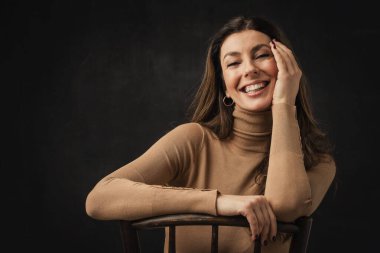 Studio portrait of an attractive middle aged woman with toothy smile wearing turtleneck sweater while sitting at isolated dark background. Copy space.