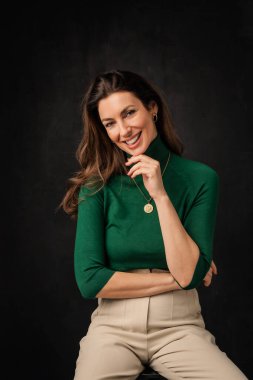 Studio portrait of an attractive middle aged woman with toothy smile wearing turtleneck sweater while sitting at isolated dark background. Copy space.