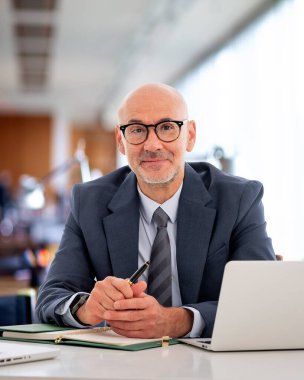 Orta yaşlı bir işadamı ofisinde oturuyor. Takım elbiseli, kravatlı ve kameraya gülümseyen kendine güvenen profesyonel bir adam..