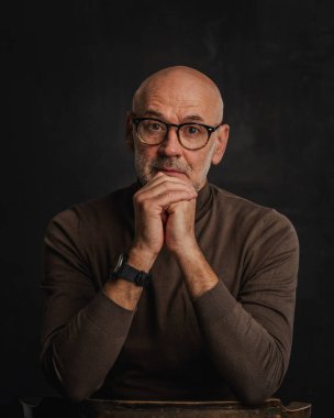 Orta yaşlı bir adam düşünceli görünüyor ve siyah arka planda oturuyor. Kel ve sakallı bir adam, izole bir arka planda günlük kıyafetler ve gözlük takıyor. Boşluğu kopyala. 