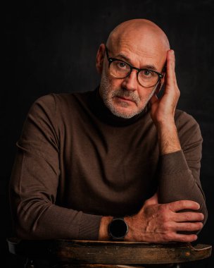 Orta yaşlı bir adam düşünceli görünüyor ve siyah arka planda oturuyor. Kel ve sakallı bir adam, izole bir arka planda günlük kıyafetler ve gözlük takıyor. Boşluğu kopyala. 