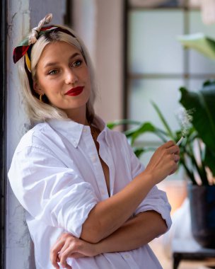 Sarışın bir kadın duvarın önünde sigara içiyor. Beyaz gömlek ve başörtüsü takan çekici bir kadın.. 