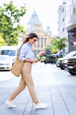 Esmer bir kadın sokakta yürüyor ve cep telefonu kullanıyor. İş günlüğü, güneş gözlüğü ve kısa mesaj takan çekici bir kadın. Tam boy çekim.