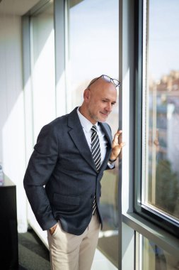 Orta yaşlı bir adam modern bir ofiste dikiliyor ve düşünceli bir şekilde pencereden dışarı bakıyor. Takım elbise ve kravat giyen kendine güvenen bir işadamı.. 