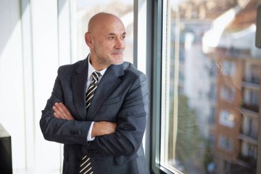 Orta yaşlı bir adam modern bir ofiste dikiliyor ve düşünceli bir şekilde pencereden dışarı bakıyor. Takım elbise ve kravat giyen kendine güvenen bir işadamı.. 