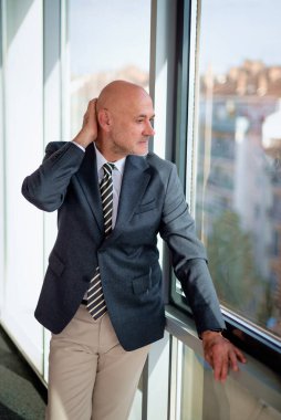Orta yaşlı bir adam modern bir ofiste dikiliyor ve düşünceli bir şekilde pencereden dışarı bakıyor. Takım elbise ve kravat giyen kendine güvenen bir işadamı.. 