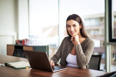 İş için bilgisayarını kullanan gülümseyen bir iş kadını. Çekici profesyonel kadın iş kıyafeti giyiyor ve modern bir ofiste oturuyor.. 