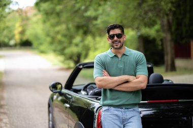 Yakışıklı, orta yaşlı bir adam üstü açık arabasının yanında duruyor ve gülüyor. Güneş gözlüğü takan ve günlük kıyafetler giyen kendine güvenen bir adam.