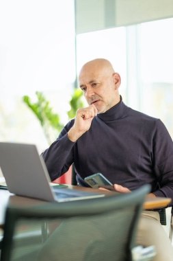 Orta yaşlı işadamı büro masasında oturuyor ve bilgisayarını kullanıyor. Profesyonel bir adam özenle bakıyor ve çalışıyor.. 