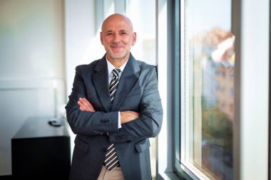 Modern bir ofiste pencerenin önünde duran orta yaşlı bir işadamı. Takım elbiseli, kravatlı ve kameraya gülümseyen kendine güvenen profesyonel bir adam..
