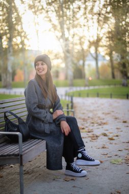 Şehir parkında oturan güzel bir kadının boyu. Gülümseyen orta yaşlı kadın palto ve bere giyiyor ve dışarıda dinleniyor..