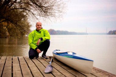 A middle-aged man sitting on the jetty and ready to go kayaking. Active sporty man wearing dry suit and looking at camera. clipart