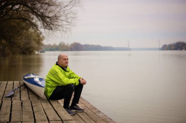 İskelede oturan ve kanoya binmeye hazır orta yaşlı bir adam. Aktif sporcu kuru takım elbise giyiyor ve başka tarafa bakıyor.. 