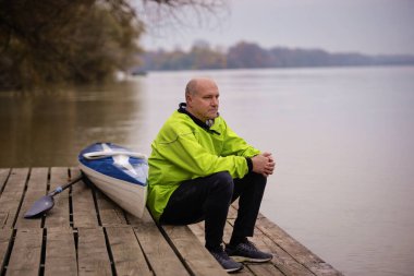 İskelede oturan ve kanoya binmeye hazır orta yaşlı bir adam. Aktif sporcu kuru takım elbise giyiyor ve başka tarafa bakıyor.. 