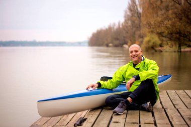 A middle-aged man sitting on the jetty and ready to go kayaking. Active sporty man wearing dry suit and looking away.  clipart