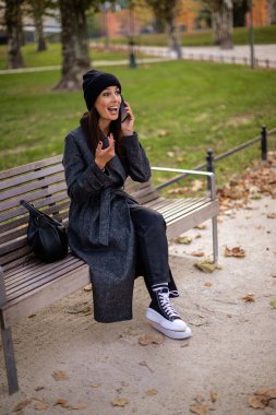 Şehir parkında oturan güzel bir kadının boyu. Güzel, orta yaşlı bir kadın ceket ve bere giyiyor ve akıllı telefonunu kullanıyor..