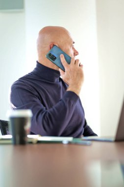 Orta yaşlı iş adamı bir dizüstü bilgisayarda çalışıyor ve modern ofis ortamında akıllı telefon kullanıyor. Gündelik kıyafetler giyen kendinden emin bir erkek.. 