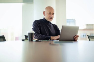 Orta yaşlı iş adamı, modern bir ofis alanında dizüstü bilgisayarda çalışıyor. Gündelik kıyafetler giyen kendinden emin bir erkek.. 