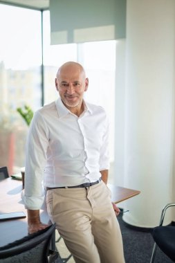 Kameraya gülümseyen orta yaşlı bir erkek. Kendine güvenen bir işadamı parlak bir ofiste duruyor..