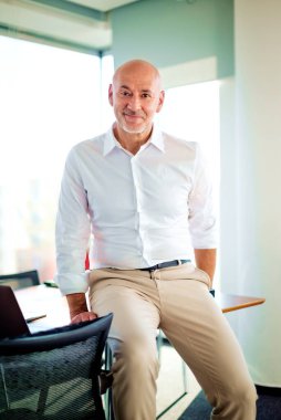 Kameraya gülümseyen orta yaşlı bir erkek. Kendine güvenen bir işadamı parlak bir ofiste duruyor..